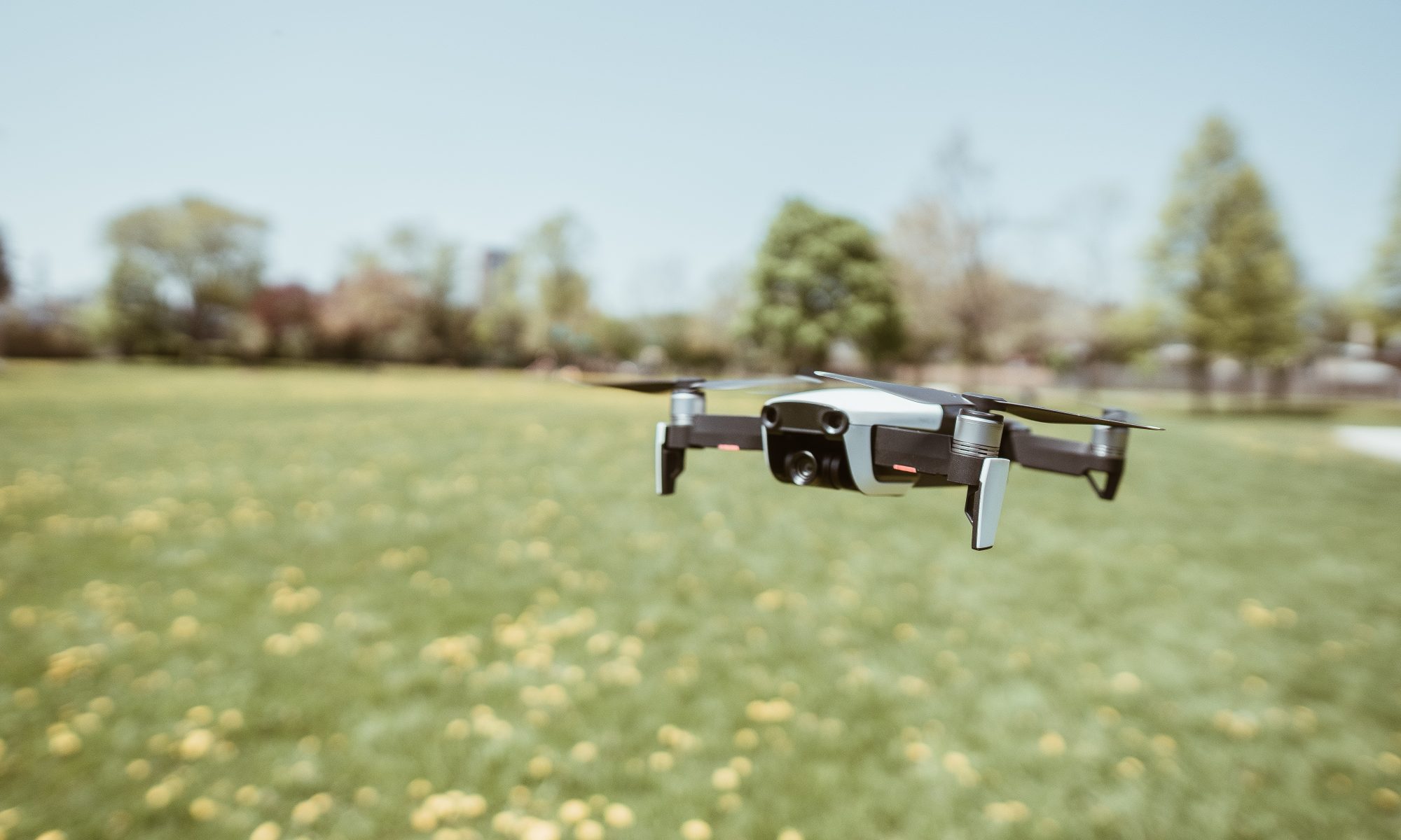 Image: Blurred background with open sky, some trees on the horizon, a field of grass and yellow flowers. A drone is hovering in the foreground.